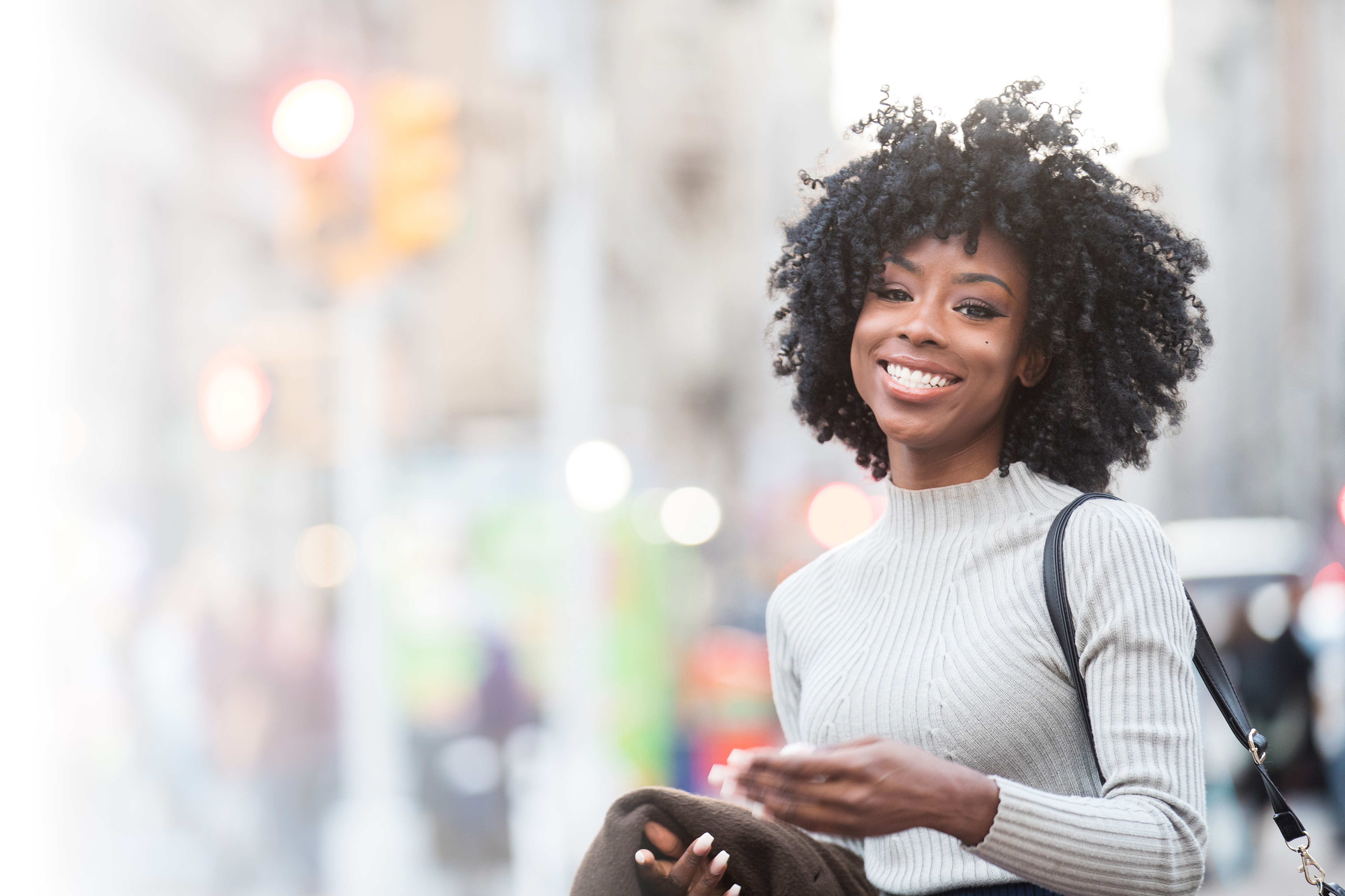 Smiling Black woman with natural curly hair wearing a light gray sweater and carrying a shoulder bag while standing on a city street.